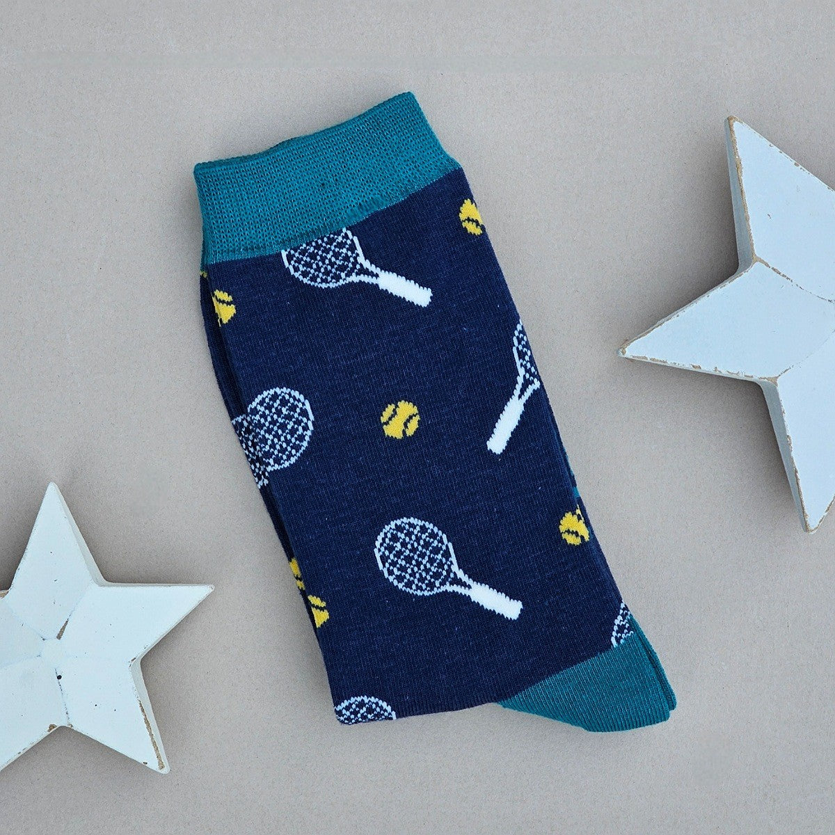 A single navy blue sock with a tennis racket and ball pattern, with a teal toe and cuff.  Styled on a neutral beige background with decorative white wooden stars.