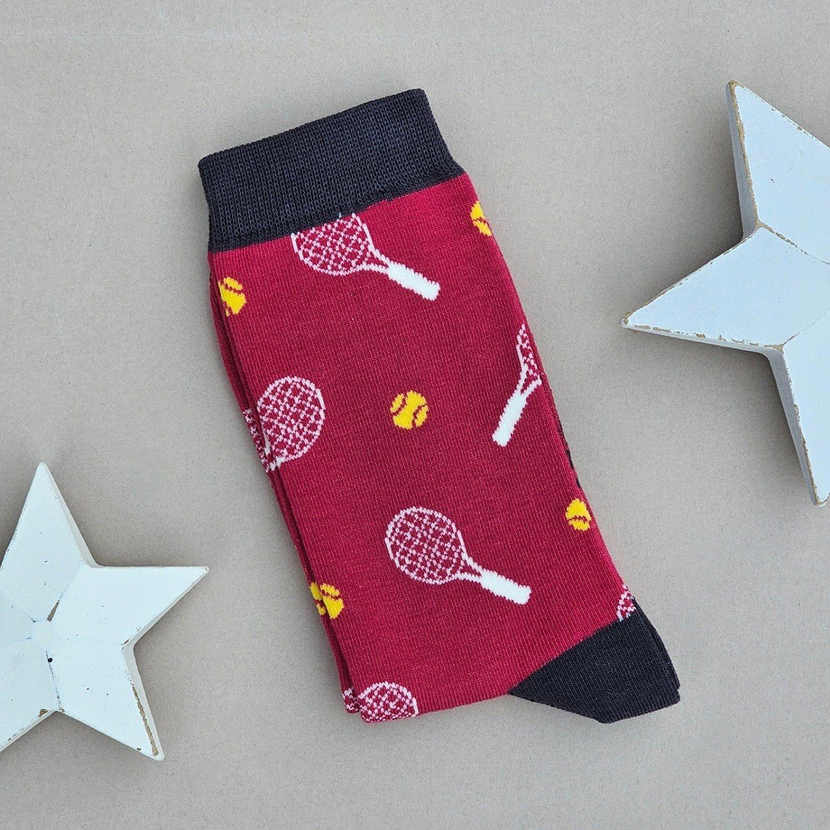 A single red sock with a tennis racket and ball pattern, with a dark grey toe and cuff.  Styled on a neutral beige background with decorative white wooden stars.