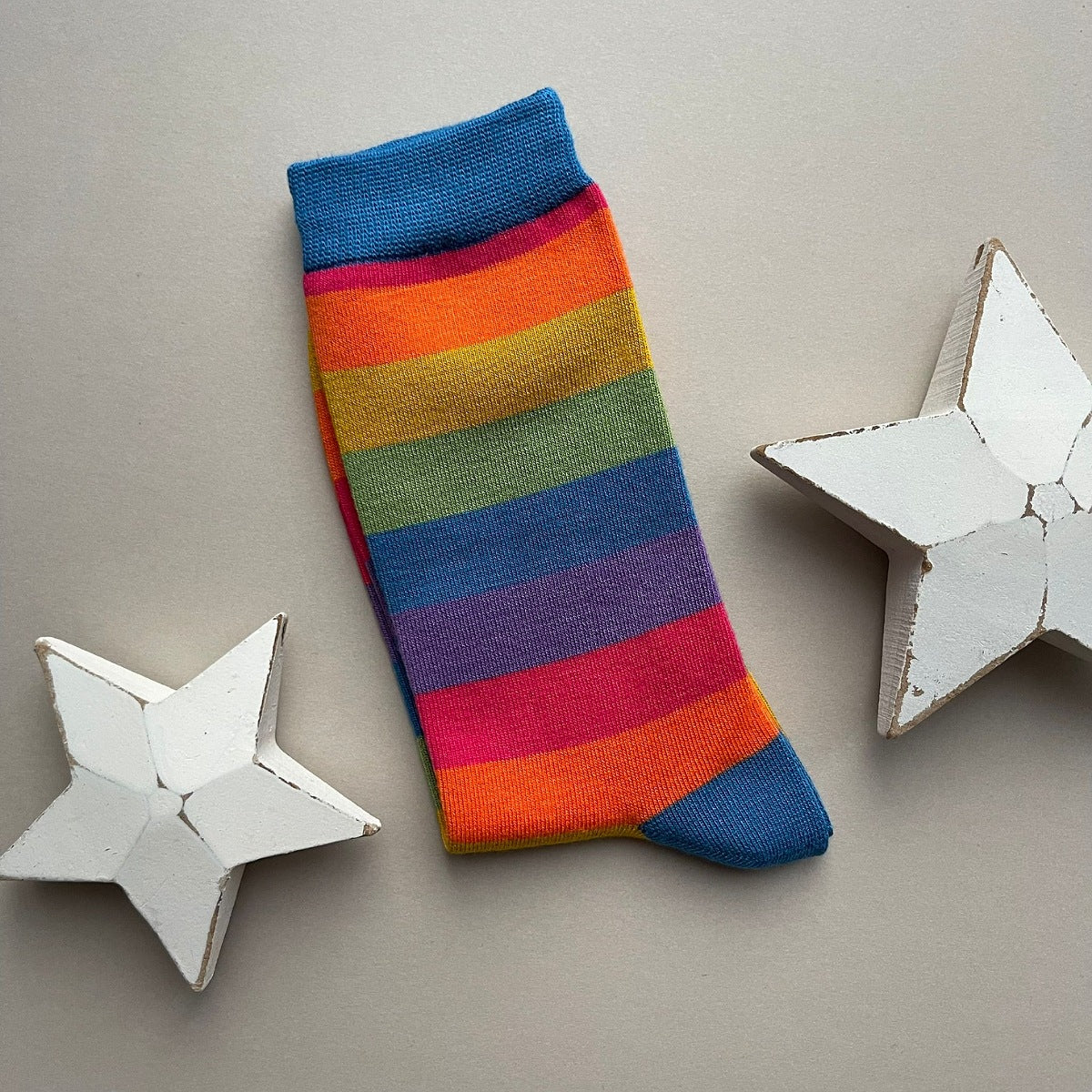 A single thick stripe sock with various colours of blue, red, orange, yellow, green and purple.
The sock is displayed on a neutral beige background with decorative white star cutouts.