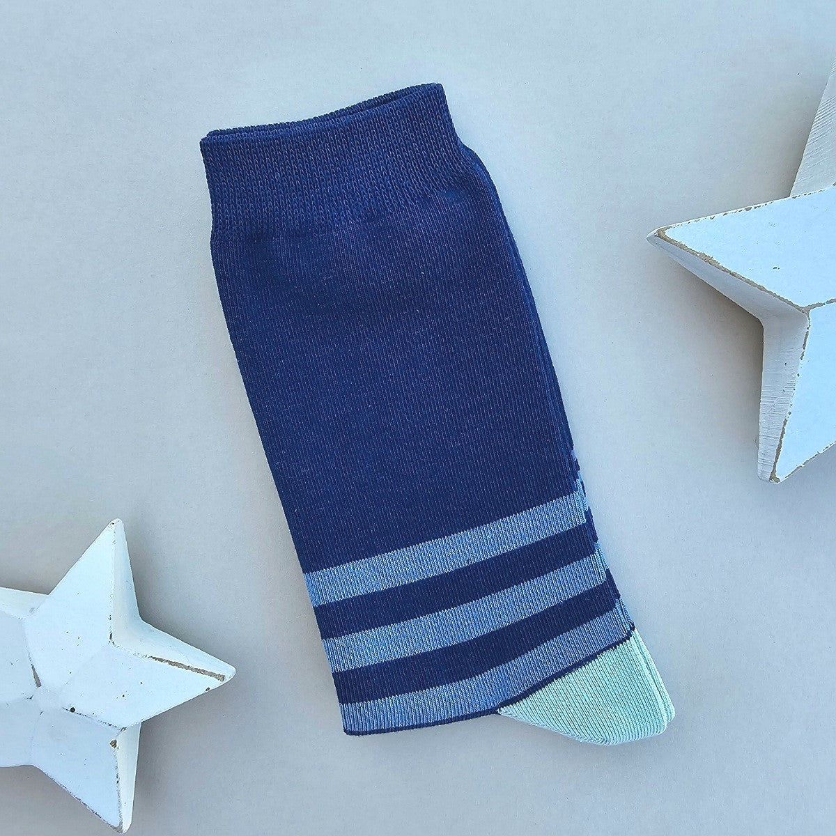 A single dark blue sock with three horizontal medium blue stripes near the toe and a light blue pointed toe cap, photographed on a pale grey background with decorative white star props.