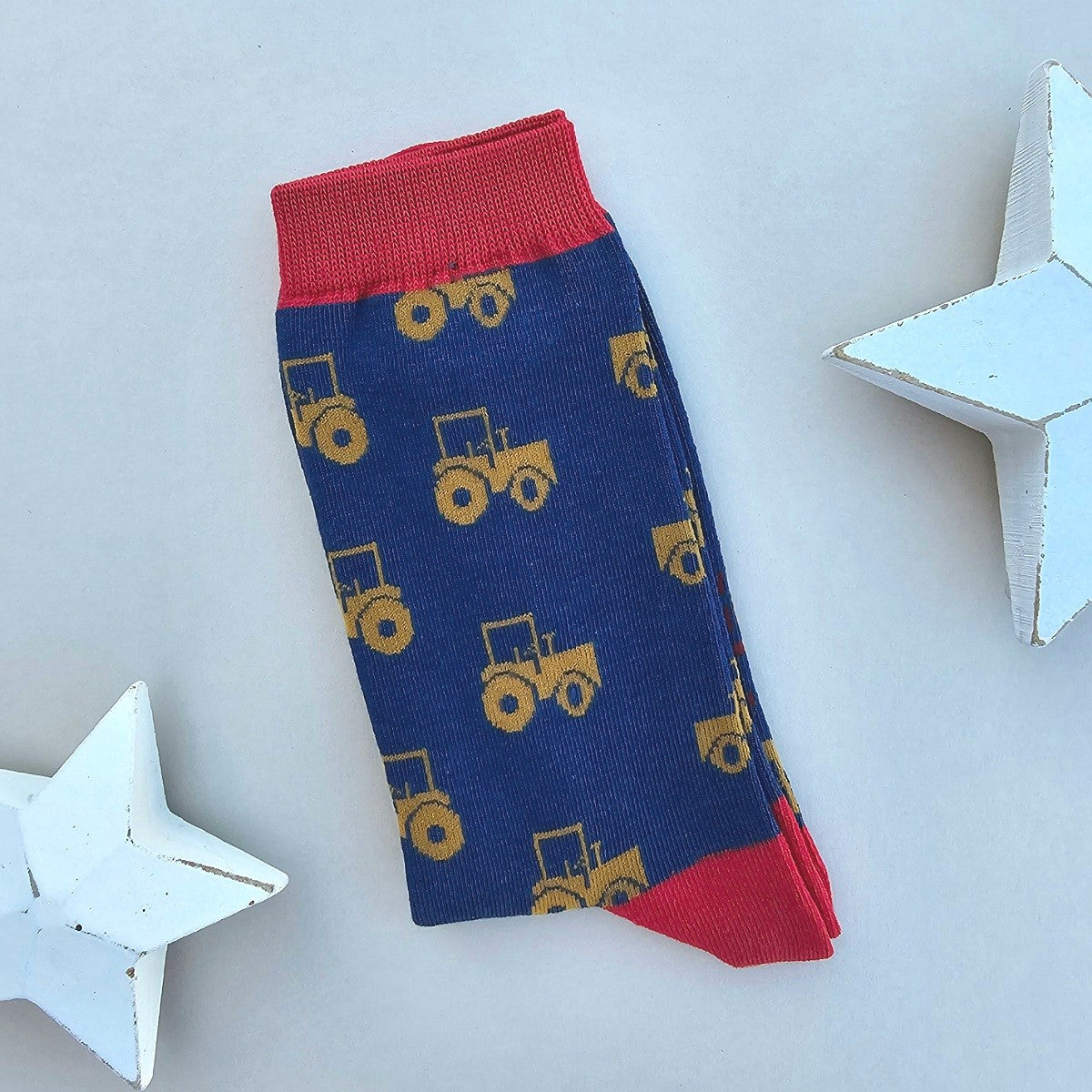 A single navy blue sock with an all-over  pattern of yellow tractors, red ribbed cuffs and heels, styled on a light grey surface with white star props.