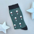 A single green sock with a pattern of white wine glasses filled with red wine, finished with black ribbed cuffs and heels, displayed on a light grey background with white star-shaped props.