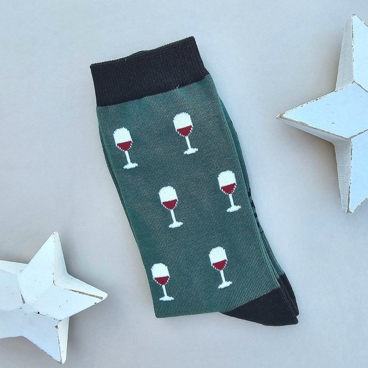 A single green sock with a pattern of white wine glasses filled with red wine, finished with black ribbed cuffs and heels, displayed on a light grey background with white star-shaped props.