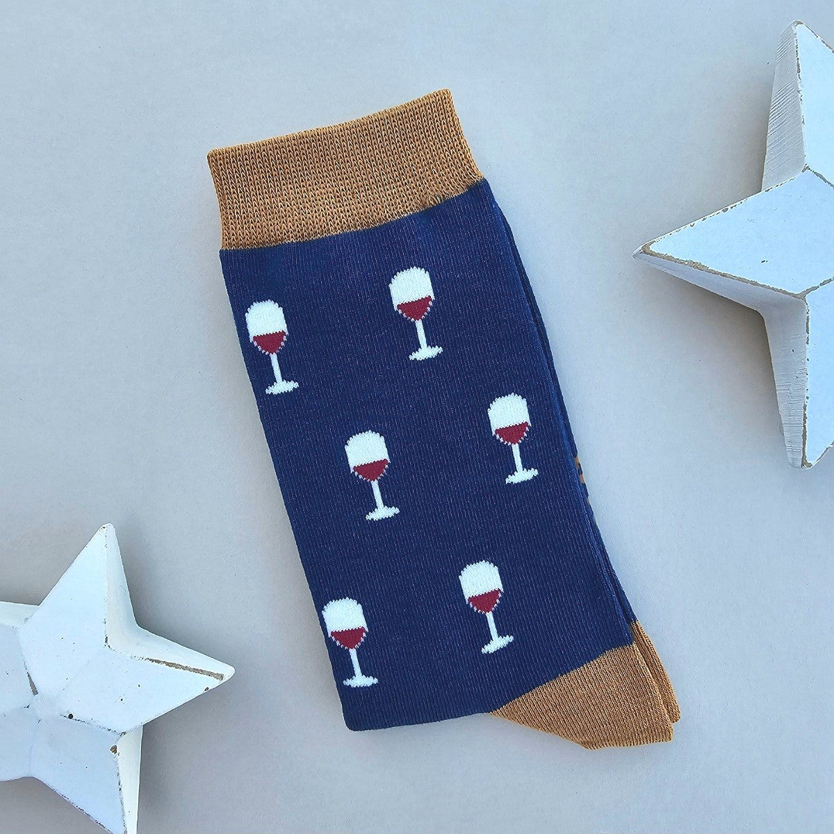 A single navy blue sock with a pattern of white wine glasses filled with red wine, finished with tan ribbed cuffs and heels, displayed on a light grey background with white star-shaped props.