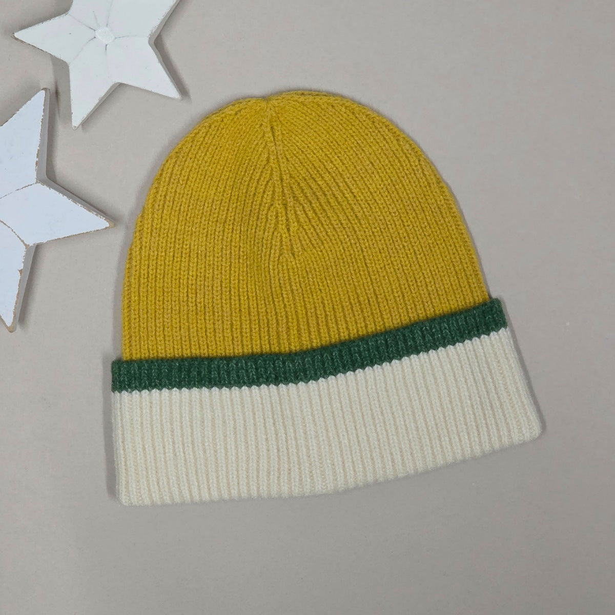 A ribbed knit beanie hat in mustard yellow with a dark green band and an off-white cuff, displayed on a neutral grey background with white star-shaped decorative props.