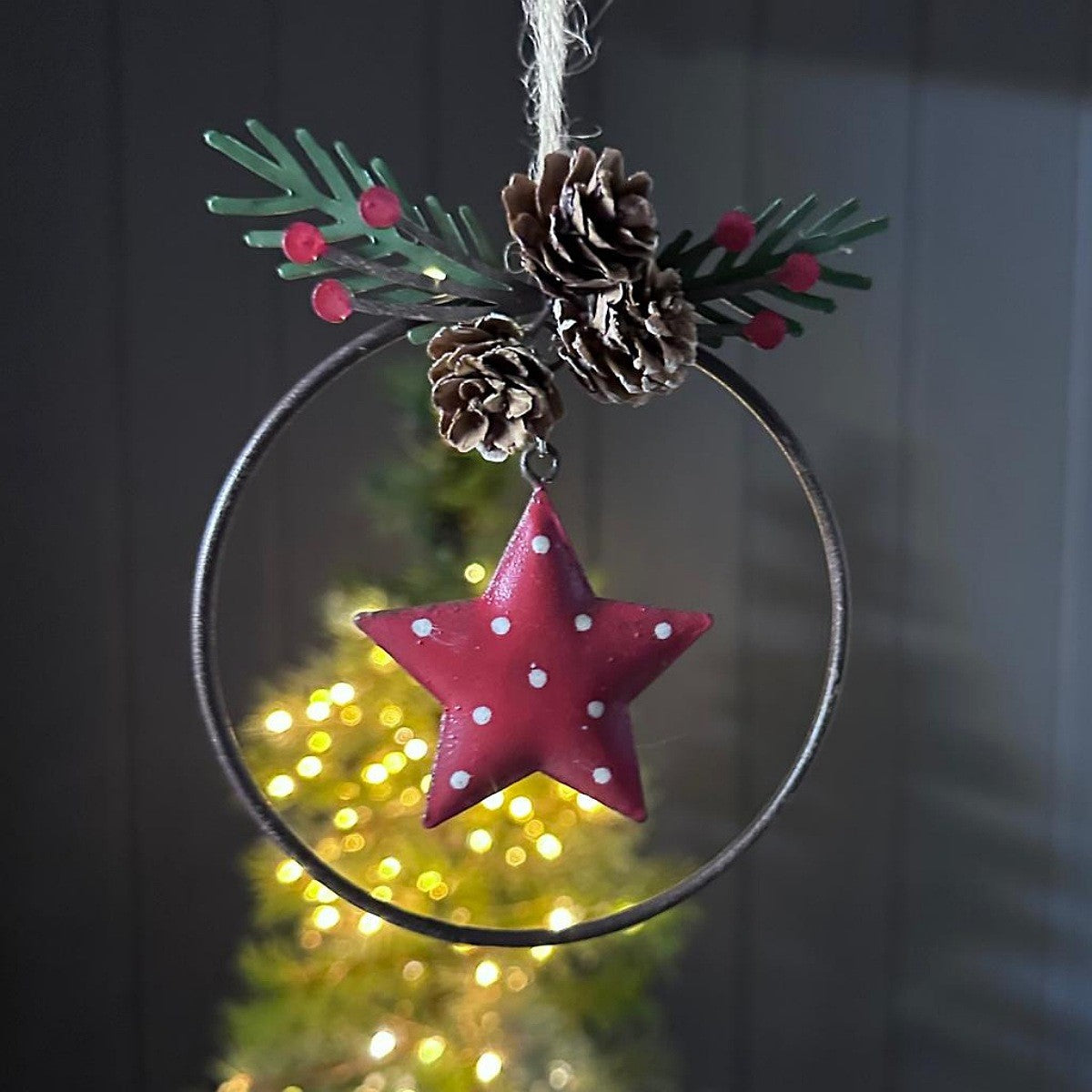 Decorative Christmas hanging ornament with a red star, pine cones, and greenery on a dark background.