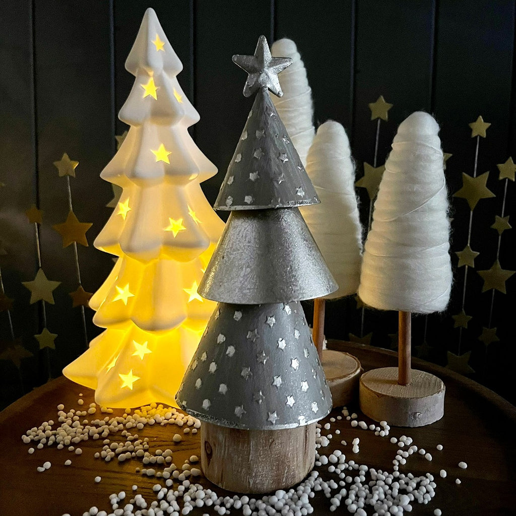 A 3 tier metallic silver tree with a star at the top, 2 tiers have mini star patterns set on a wooden base.  Set in-front of  christmas tree ornaments displayed on a wooden surface against a dark panelled background and a few gold star-shape in the background.