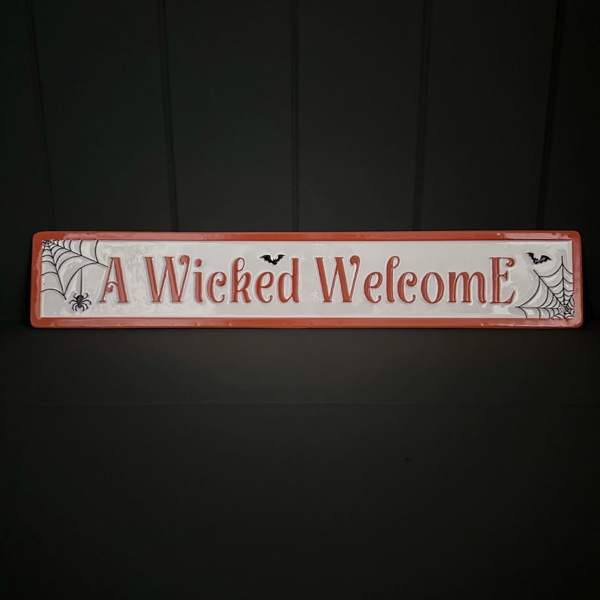 A rectangular Halloween sign with a distressed white background, orange border, and raised orange text reading 'A Wicked Welcome.' Decorative black spiderwebs, spiders, and bats appear on the sign.