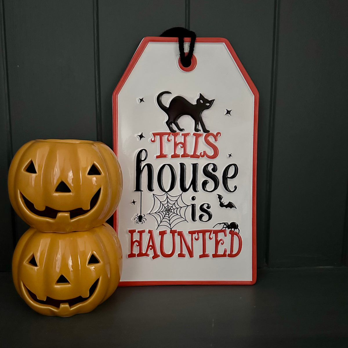 A Halloween decorative sign that reads 'THIS house is HAUNTED' in red and black lettering on a white background with a red border, shown next to two small orange jack-o'-lantern figurines against a dark paneled backdrop.