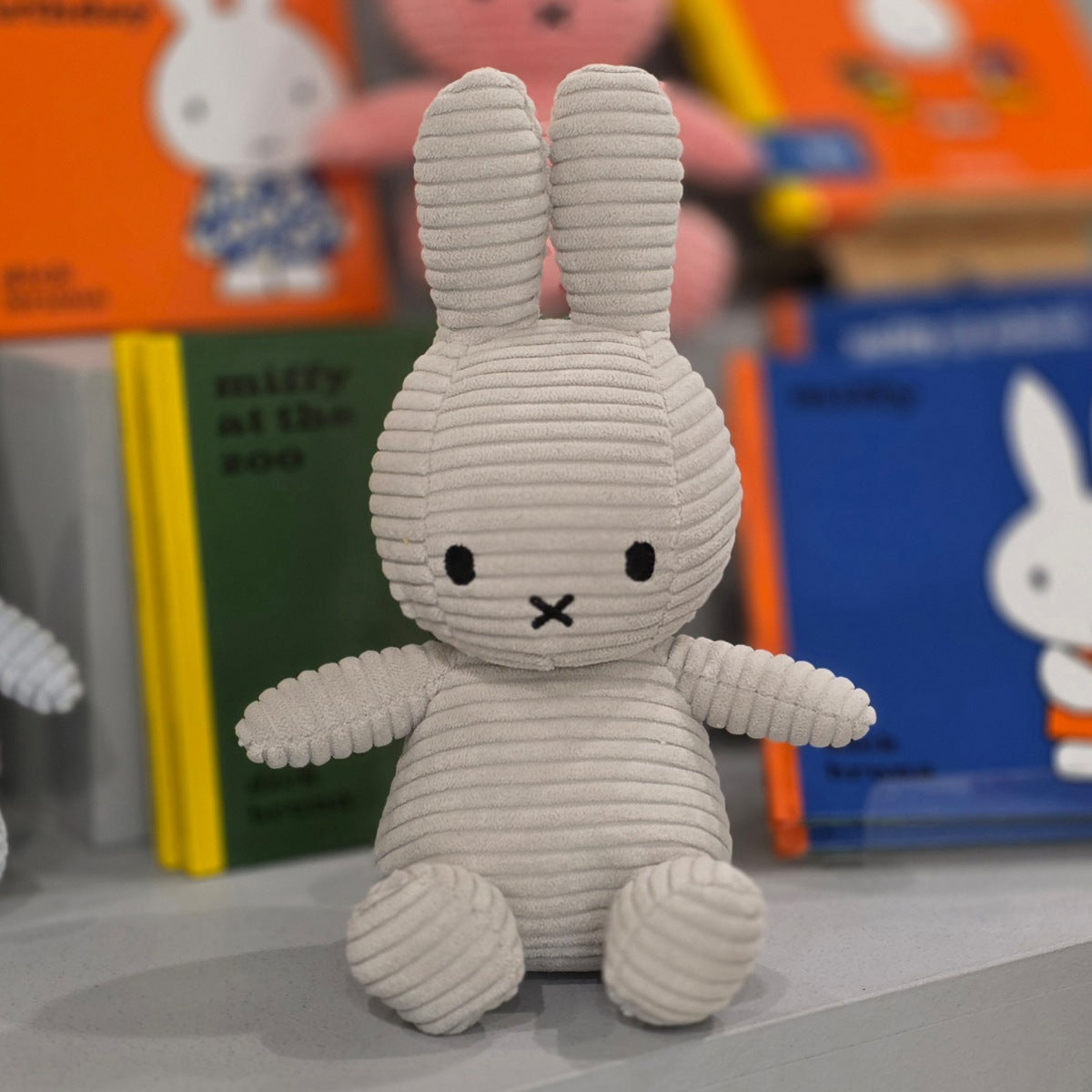 A plush rabbit toy made of ribbed corduroy fabric, styled like Miffy, sitting in front of colorful children's books on a shelf.