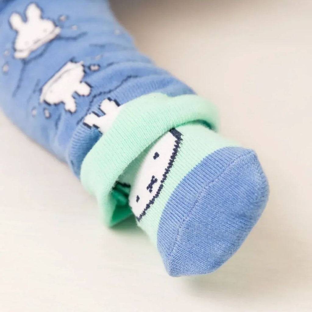 A close-up of a folded pair of baby socks with a light blue body, mint green cuff, and white cartoon Miffy bunny pattern, displayed on a light beige surface.