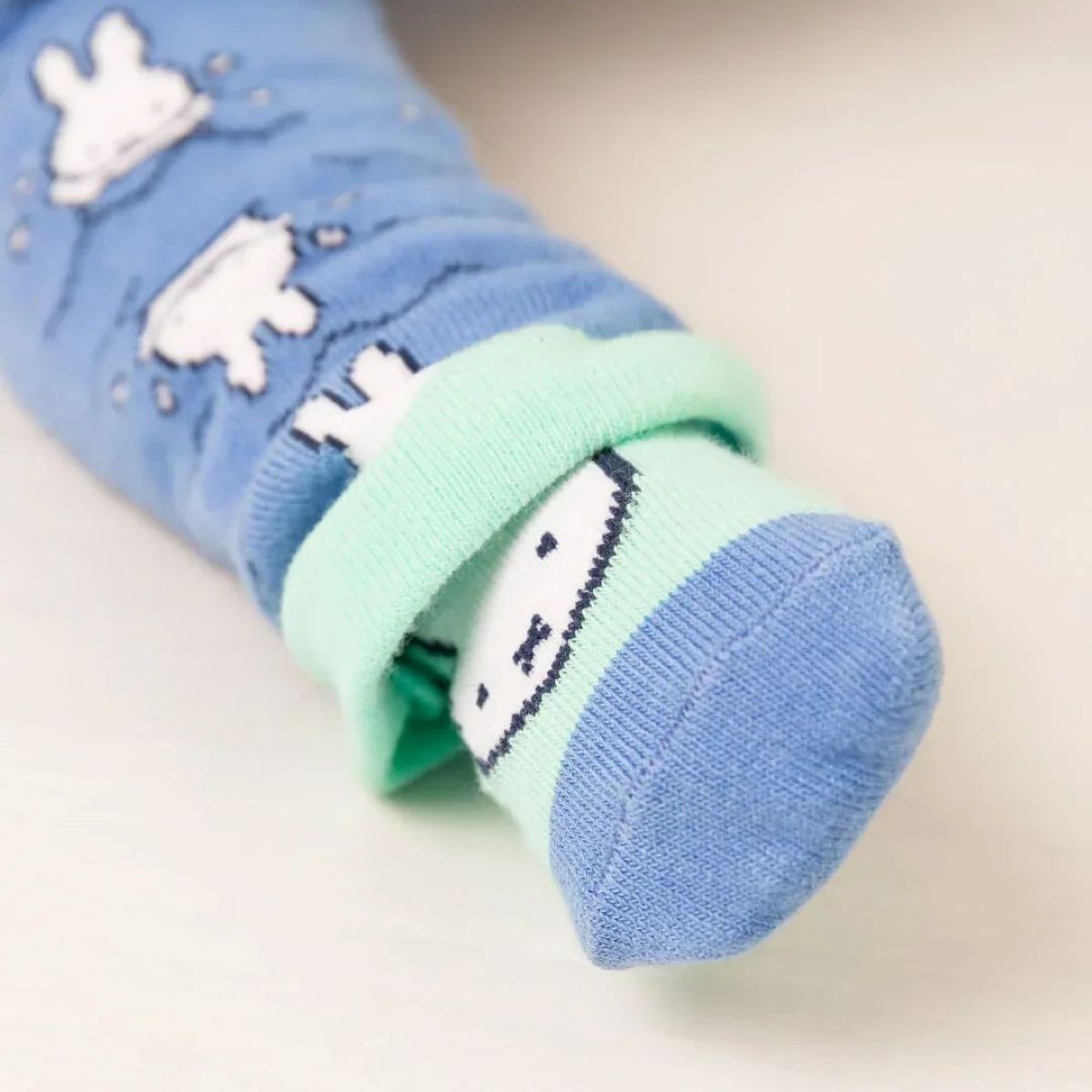 A close-up of a folded pair of baby socks with a light blue body, mint green cuff, and white cartoon Miffy bunny pattern, displayed on a light beige surface.