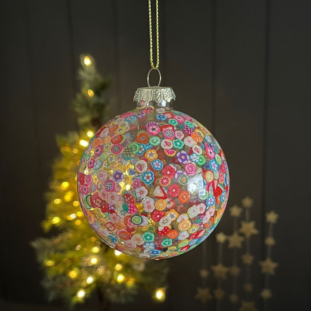 A clear glass Christmas ornament ball covered in colorful button-like patterns, hanging from a gold string against a dark background with a blurred lit Christmas tree.
