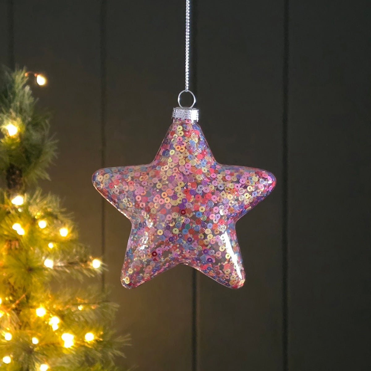 A five-pointed star-shaped Christmas ornament covered in iridescent tiny sequins, hanging from a silver string against a dark panelled background with a softly blurred lit Christmas tree on the left.