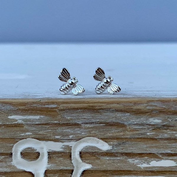 A pair of small silver bee stud earrings resting on a weathered wooden surface with a blurred blue-gray background.
