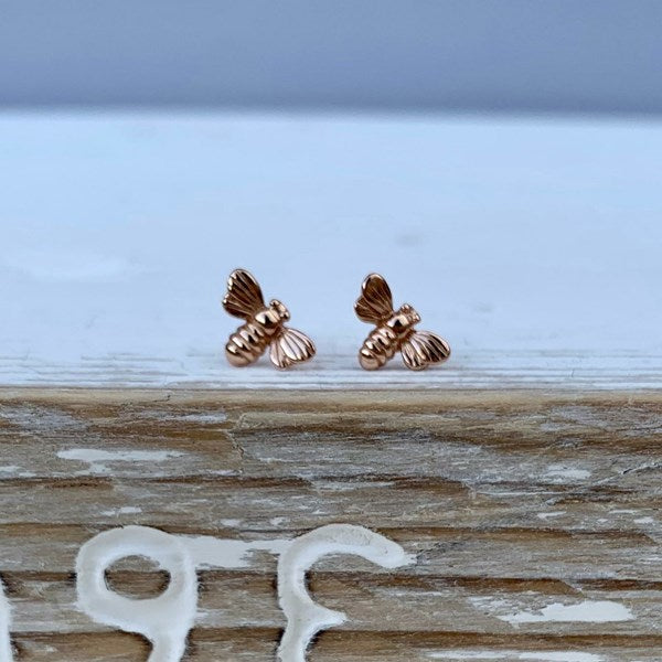 A pair of rose gold bee stud earrings resting on a rustic wooden surface with a light blue background.