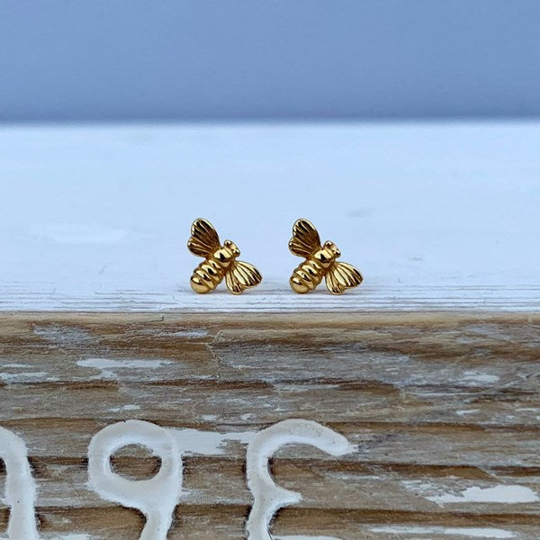 A pair of small gold bee stud earrings resting on a weathered wooden surface with a soft blue-gray background.