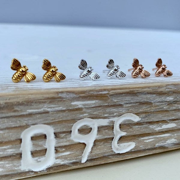 Set of bee-shaped earrings in gold, silver, and rose gold on a wooden block.