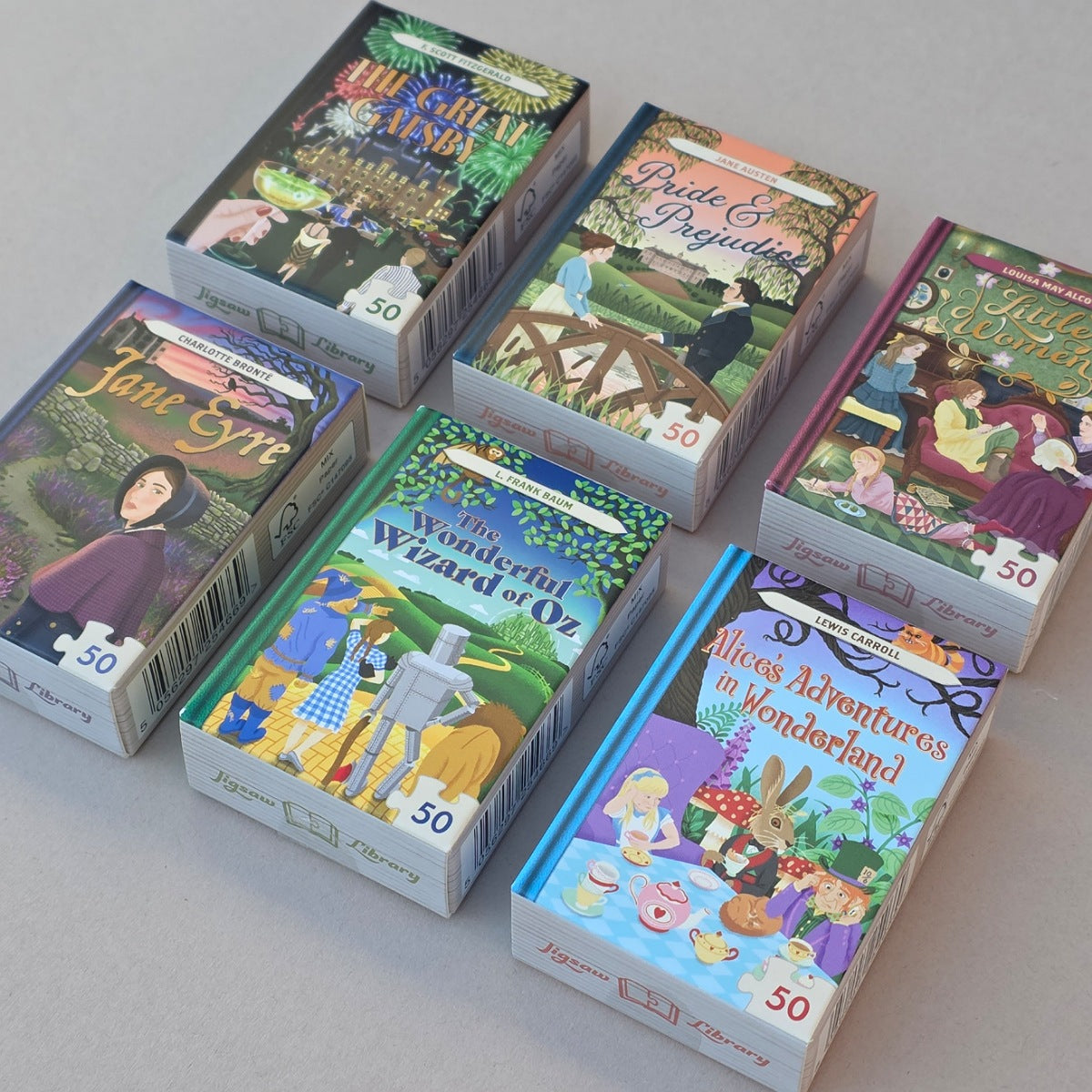 Six illustrated jigsaw puzzle boxes arranged on a light grey surface. The boxes feature colorful vintage-style covers with puzzle imagery and titles including Jane Sykes, The Wizard of Oz, Alice Adventures in Wonderland, and others.