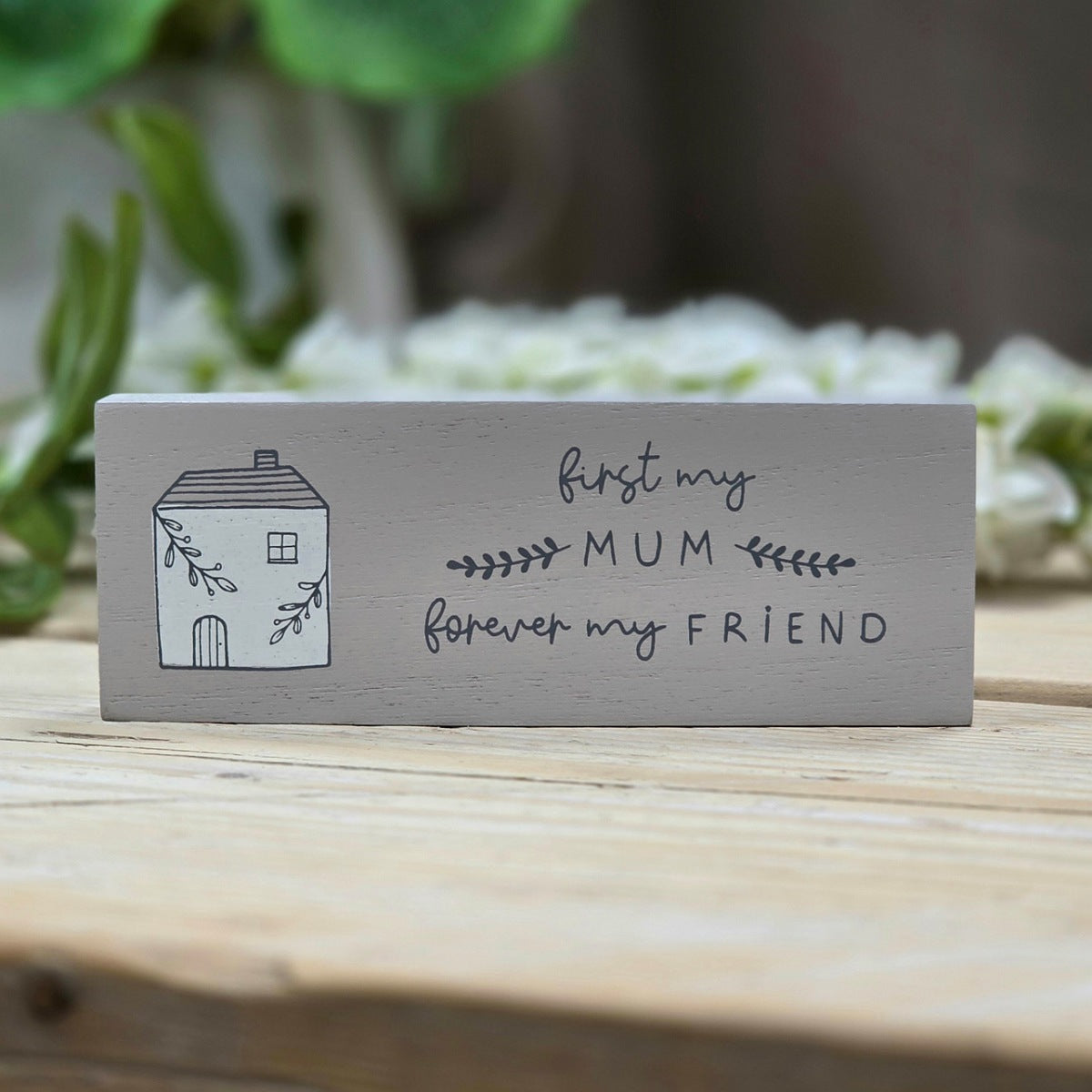 A rectangular light grey wooden sign with an image of a white house the phrase 'first my MUM forever my FRIEND' in dark script and a small house illustration, displayed on a wooden surface with a blurred garden background.