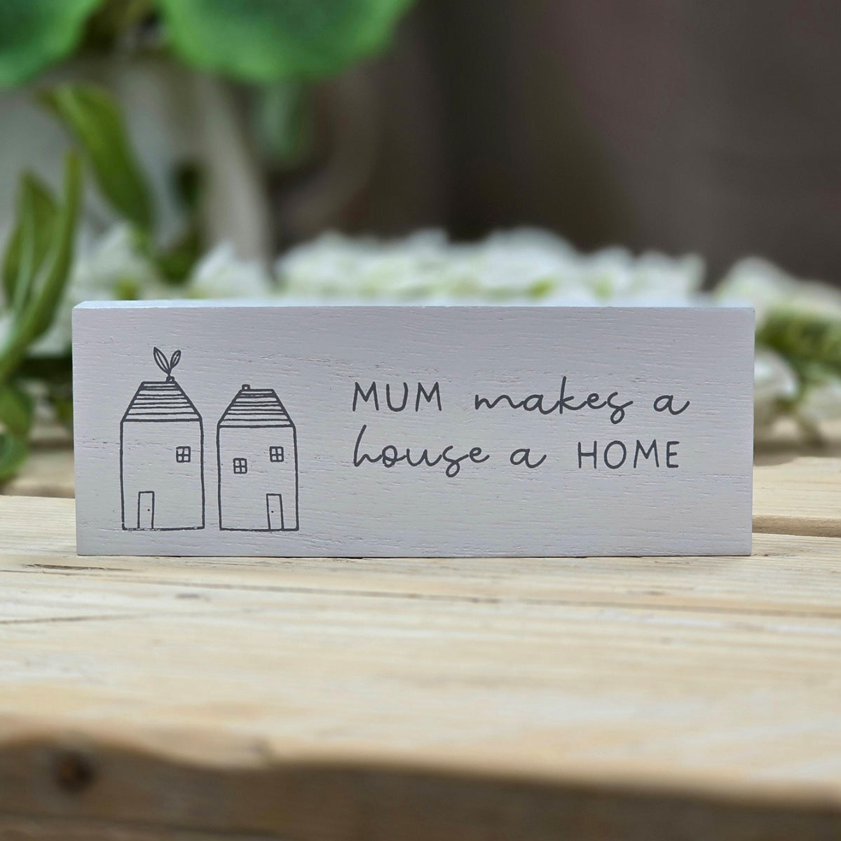 A rectangular white wooden sign with a hand-painted black design of two small houses and the phrase 'MUM makes a house a HOME', displayed on a light wooden surface with a blurred garden background.