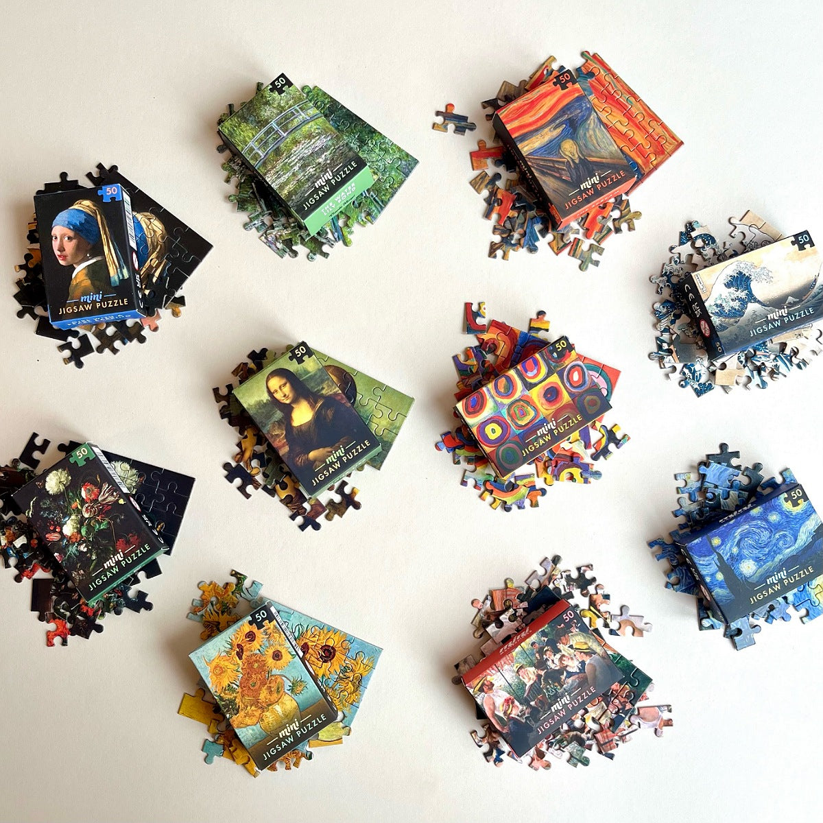 A flat-lay arrangement of matchbox sized jigsaw puzzle boxes and scattered puzzle pieces on a light  background. The boxes feature famous artwork and are labeled with artist names such as Pablo Picasso, Mona Lisa, Van Gogh, Da Vinci, and Starry Night.