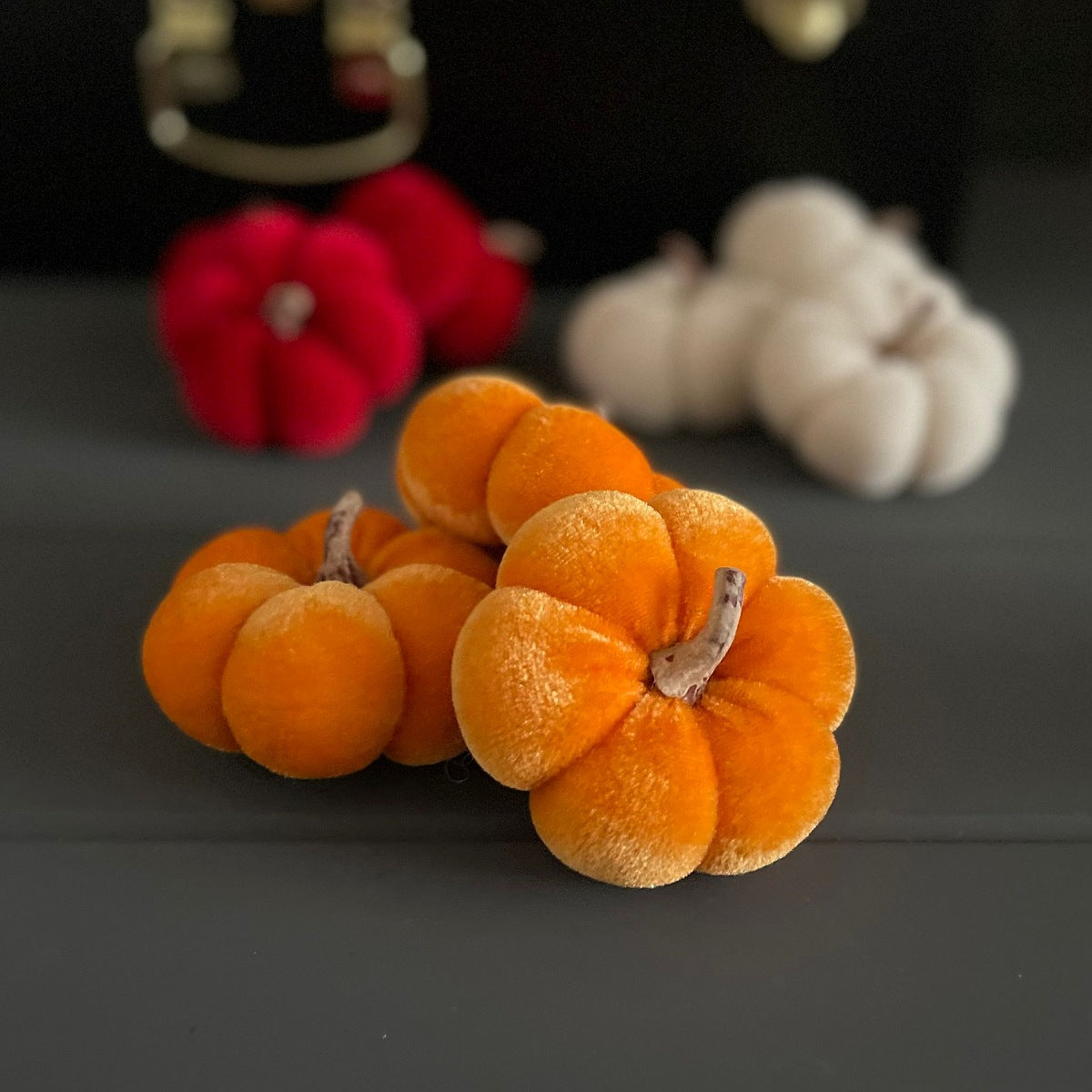 Three small plush velvet pumpkins in orange, red, and off-white, arranged on a dark grey surface with a blurred black background.