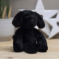 A black plush dog toy with floppy ears sits on a light wood surface, with a blurred white star decoration and a plant in the background.