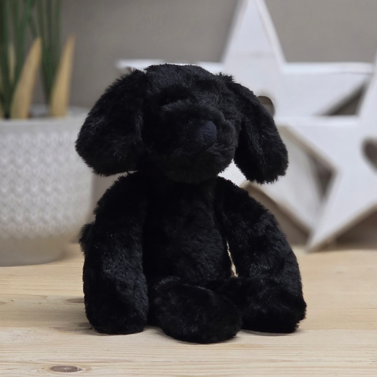 A black plush dog toy with floppy ears sits on a light wood surface, with a blurred white star decoration and a plant in the background.