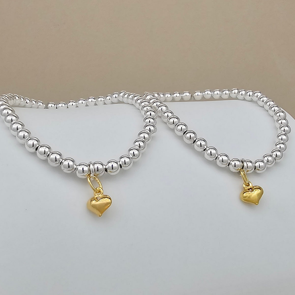 Two beaded bracelets with polished silver-toned round beads and small dangling gold heart charms, displayed on a light surface against a beige background.