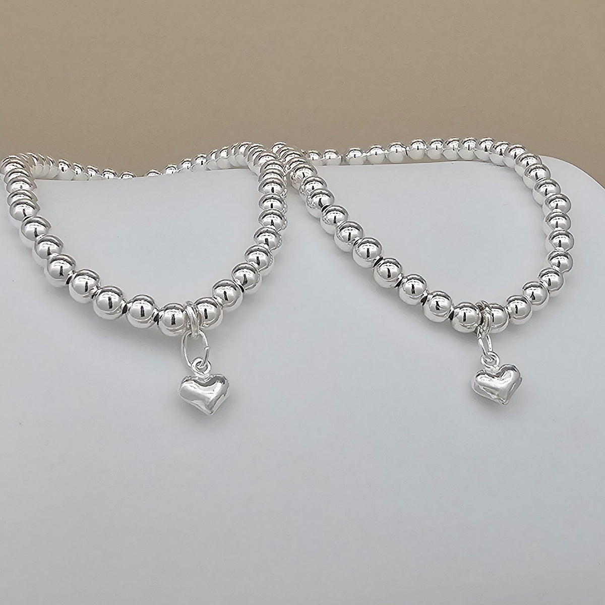 Two silver beaded bracelets with small heart charms, displayed on a white surface against a beige background.
