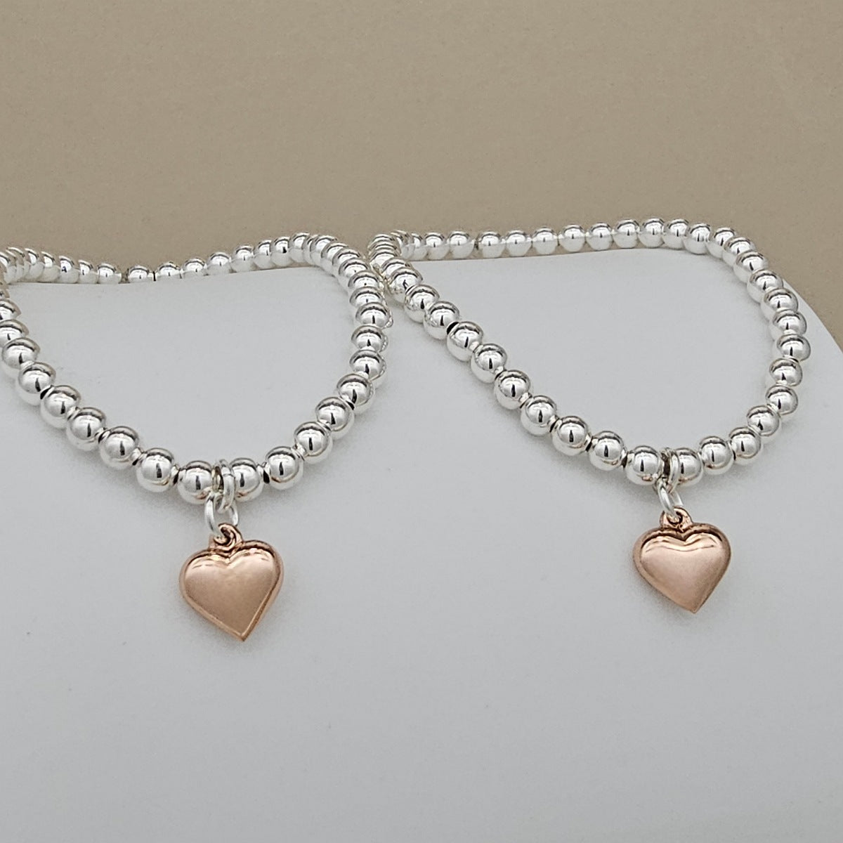 Two silver beaded bracelets with rose gold heart charms on a light gray background