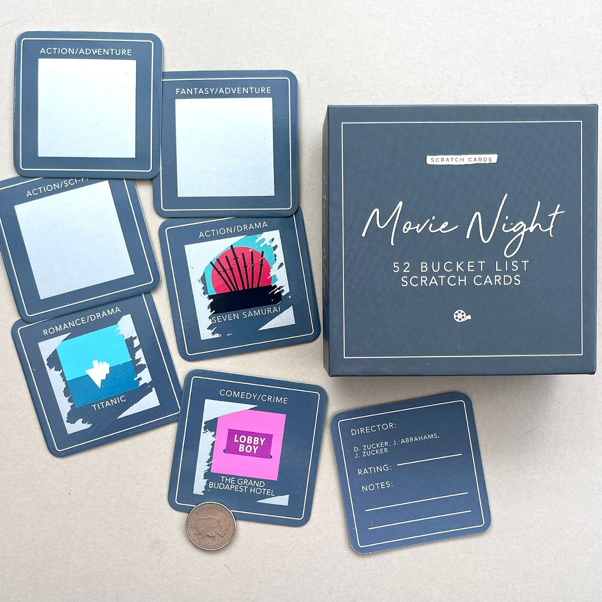 Movie Night Scratching Cards set with a grey box and several sample cards laid out on a light beige surface, including card categories like Action/Adventure, Fantasy/Adventure, Romance/Drama, Comedy/Crime, and a Director card, with a penny for scale.