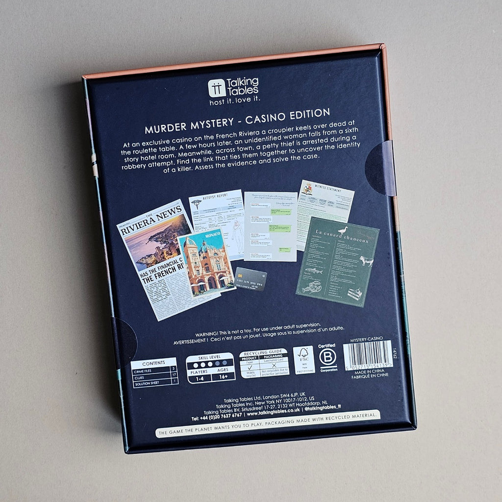 The back of the 'Murder Mystery - Casino Edition' board game box, photographed from above on a neutral gray surface. The box is dark navy blue with rose gold trim along the top edge and a side tab.
