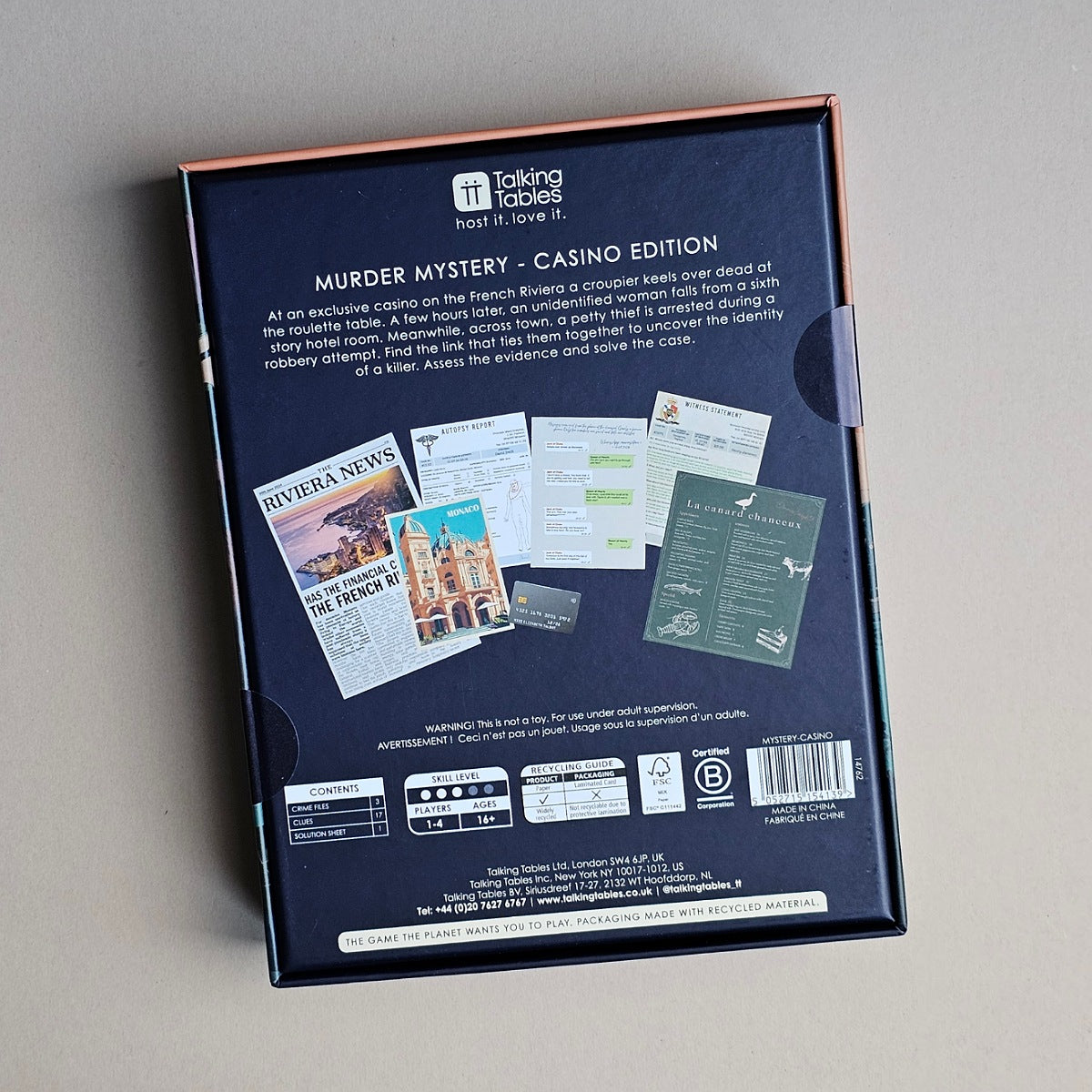 The back of the 'Murder Mystery - Casino Edition' board game box, photographed from above on a neutral gray surface. The box is dark navy blue with rose gold trim along the top edge and a side tab.