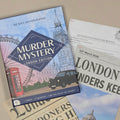A boxed game or puzzle titled "Murder Mystery London Edition" lies on top of printed case-files and newspaper clippings. The box features illustrated London landmarks, a red phone, a black cab, and the text "Deadly Destinations" and "Murder Mystery London Edition.