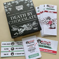 A tabletop flat-lay of a Murder Mystery Party board game set titled 'Death by Chocolate.' The black game box is shown with several white player cards, evidence cards, and a deck of chocolate-themed cards arranged on a beige surface.