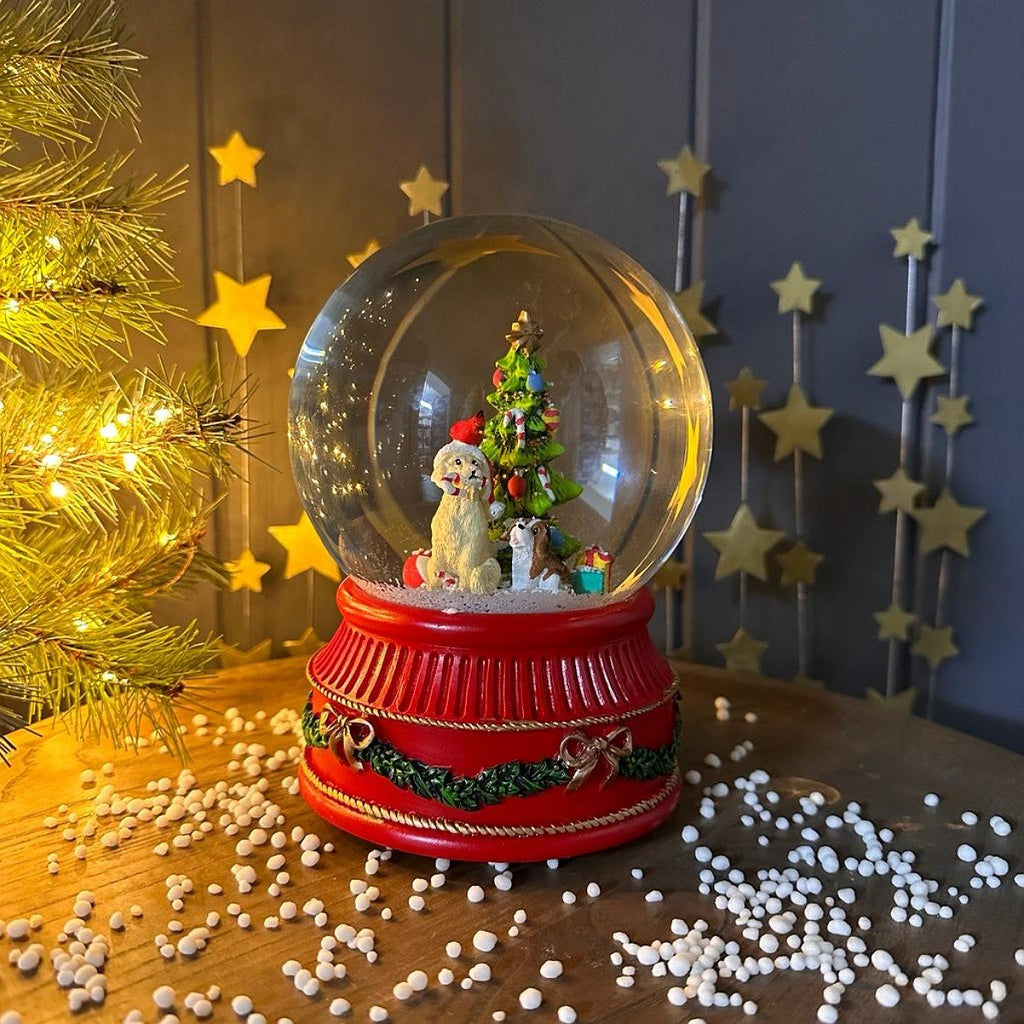A Christmas-themed snow globe with a red base, decorated with a miniature Christmas tree, gift, and two small dog figurines. The globe sits on a wooden table scattered with white beads, with a lit gold Christmas tree on the left and a dark gray paneled wall with hanging gold stars in the background.
