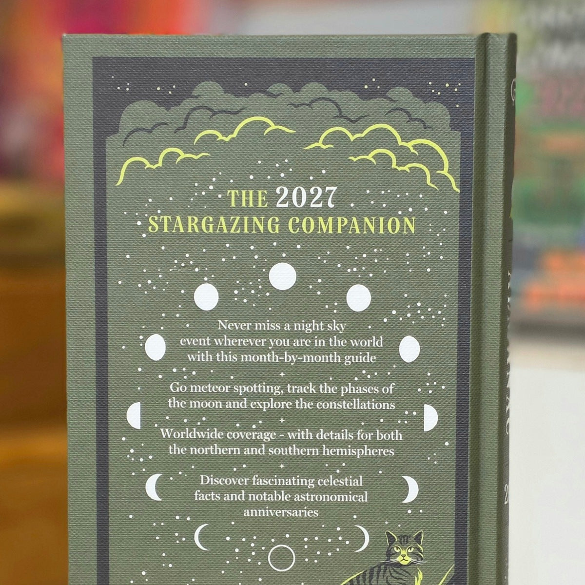 The cover of a hardcover book titled 'The 2027 Stargazing Companion' is shown upright. The cover is dark green with a starry night sky design, yellow clouds, white moon phases, and gold text.