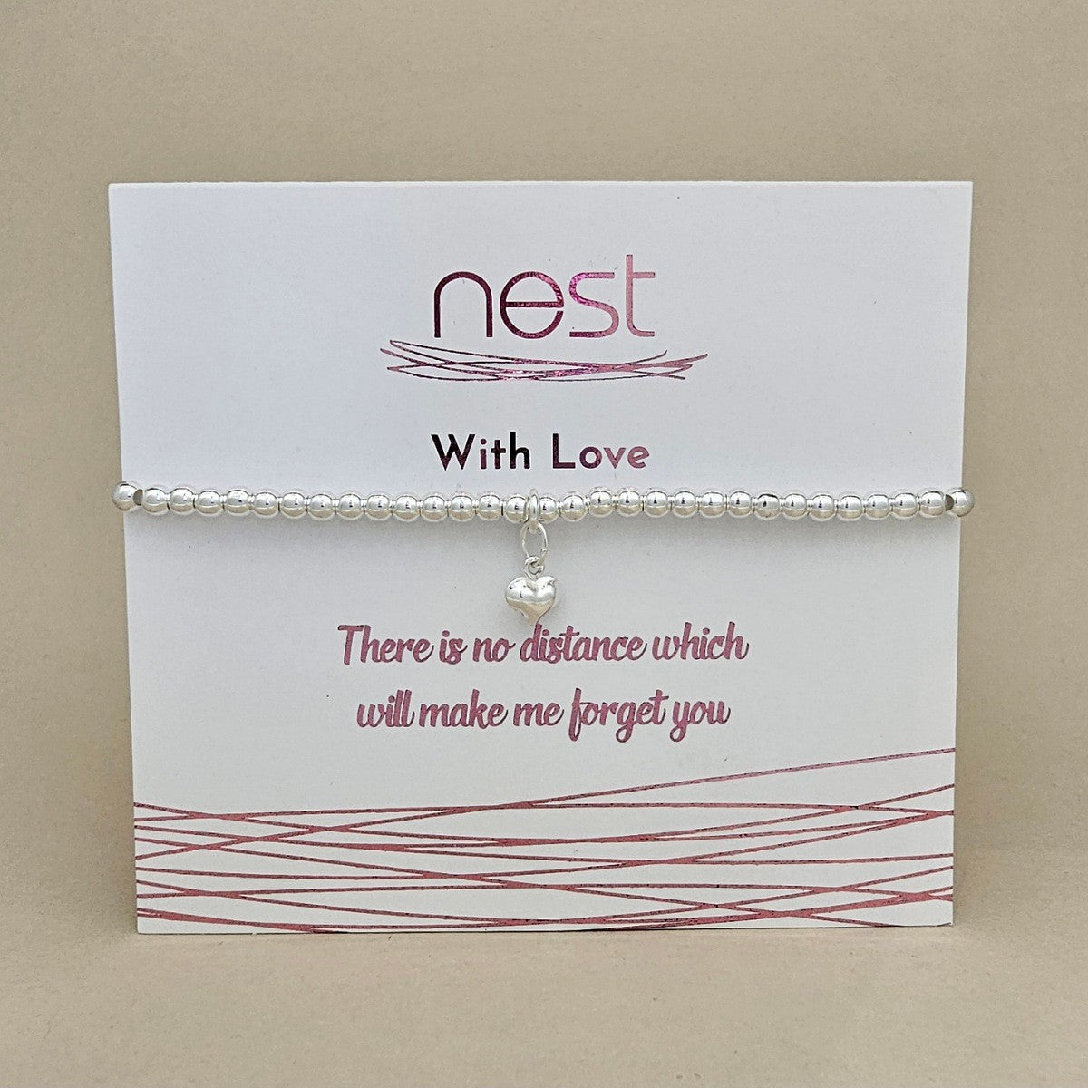 A silver beaded bracelet with a small pearl-like heart charm, displayed on a white card from the brand nest. The card features pink text reading 'With Love' and a sentimental quote, set against a solid beige background.
