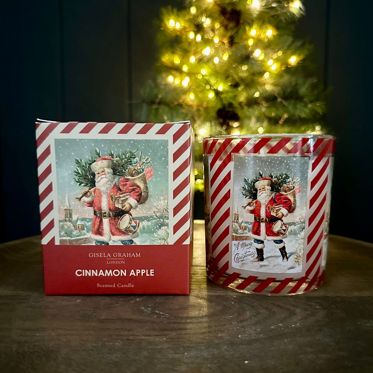 A Gisela Graham scented candle in a red and white striped glass jar next to its matching retail box, featuring a vintage illustration of Santa Claus, set on a wooden table with a blurred Christmas tree in the background.