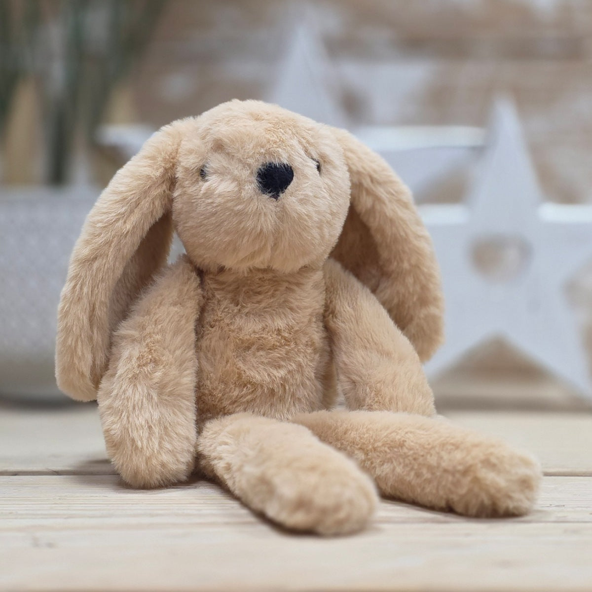 A plush stuffed bunny in a light tan color, sitting on a light wooden surface with a softly blurred background featuring a white star decoration.
