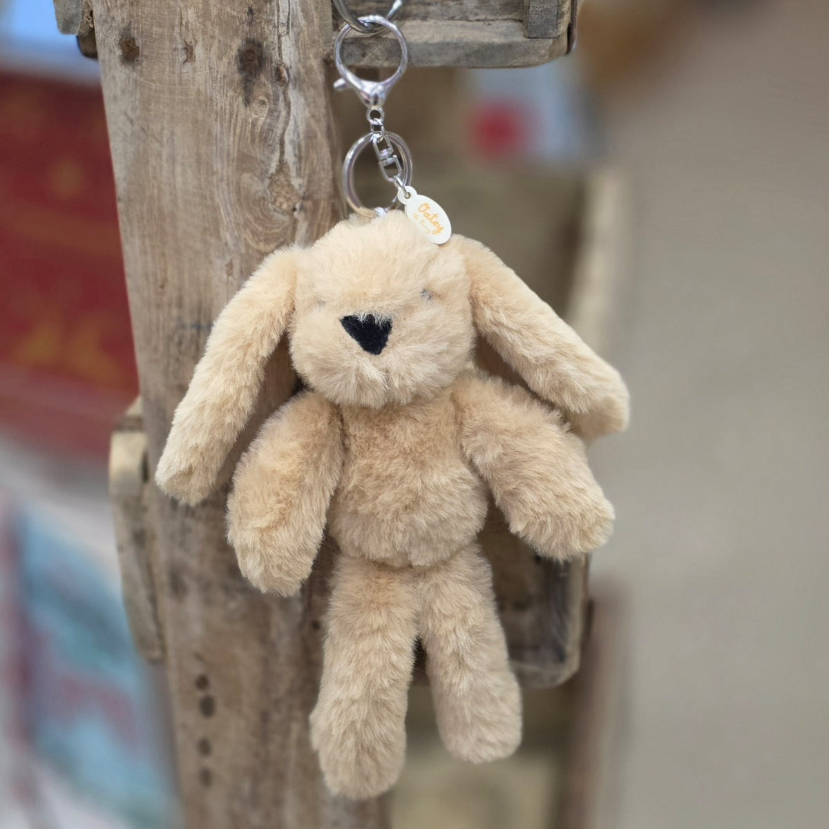 A light brown plush bunny keychain hanging from a rustic wooden post. The keychain features a soft, fuzzy light brown bunny with black eyes and nose, attached to a silver metal key ring and a small round tag.
