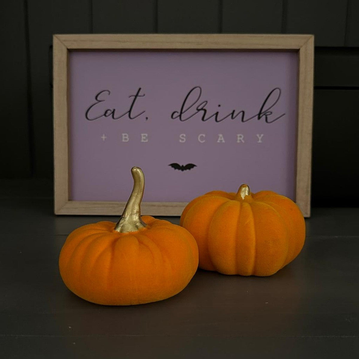 A framed decorative sign reading 'Eat, drink + BE SCARY' in black script and block lettering on a lavender background, styled with two small velvet orange pumpkins in front on a dark surface.