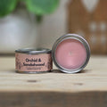 An open round metal candle tin with pink wax sits beside a closed matching tin on a wooden surface. The label reads 'Orchid & Sandalwood' and 'Scent Palette.