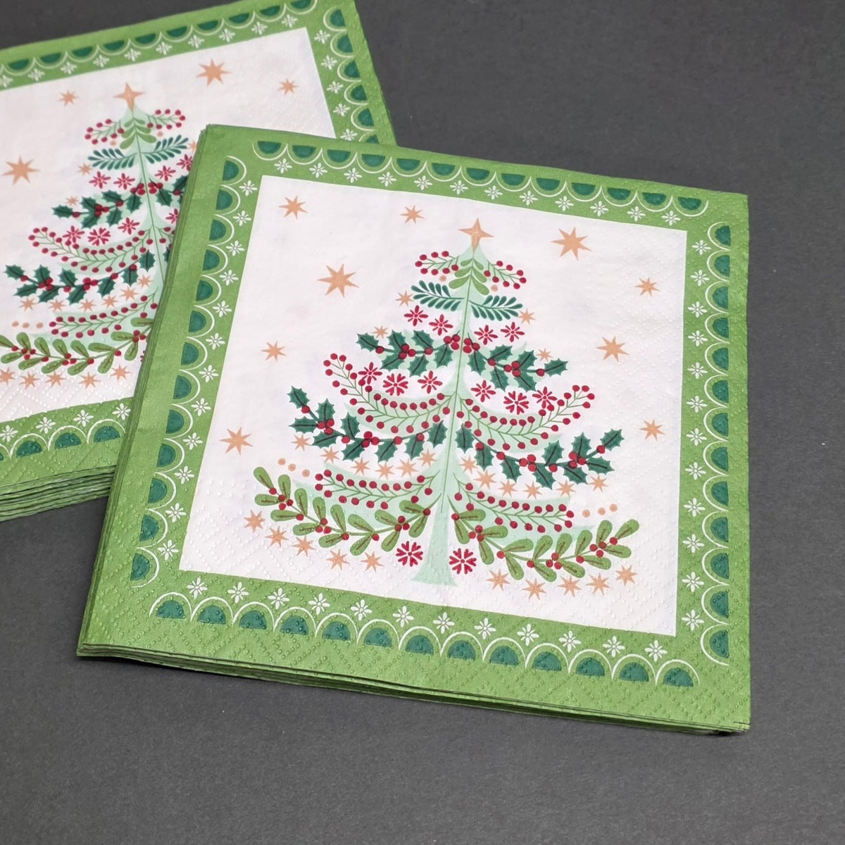 A set of square festive napkins featuring a Christmas tree design with green holly, red berries, and gold stars, displayed on a dark grey surface.