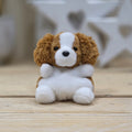 A small plush dog toy with brown and white fur, sitting on a light wooden surface with a softly blurred white decorative background.