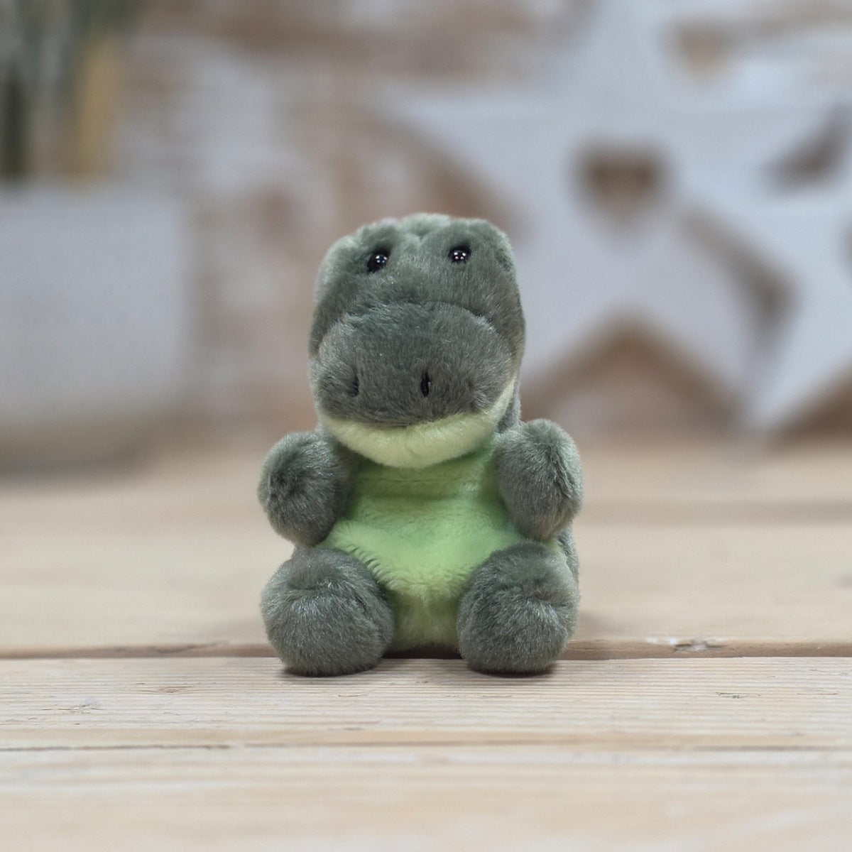 A small plush dinosaur toy with a dark gray body, light green belly, and cream-colored under-chin, sitting on a light wooden surface with a blurred background.