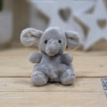A small plush elephant toy in light gray sits centered on a light wood surface with a softly blurred indoor background.