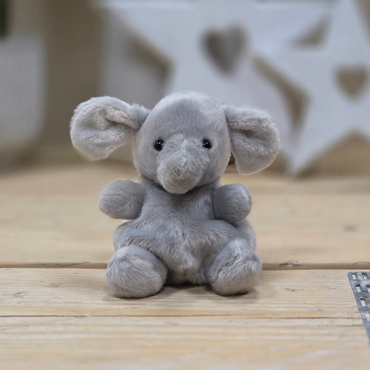 A small plush elephant toy in light gray sits centered on a light wood surface with a softly blurred indoor background.